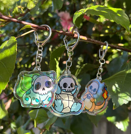 An acrylic keychain with double sided printing of Bulbasaur, Charmander and Squirtle 'Skelemon' design. Back side has a holographic shards finish.